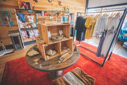 A cozy boutique interior featuring a wooden display table, clothing racks, and colorful decor on a vibrant rug.