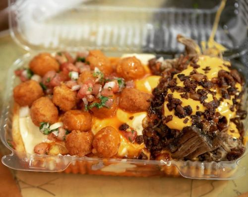 A takeout container filled with cheesy nachos, tater tots, and pulled meat topped with bacon bits and sauce.