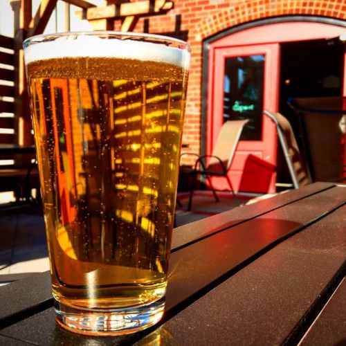 A glass of golden beer sits on a table, reflecting sunlight and the surrounding outdoor setting.