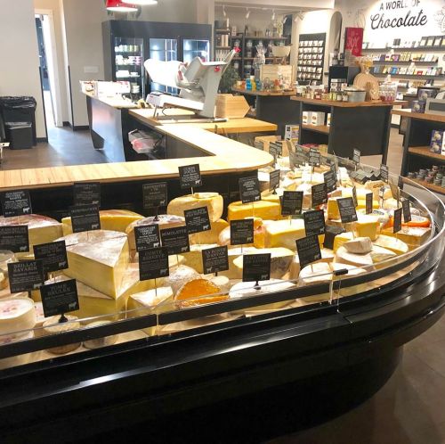 A display case filled with various cheeses, labeled with signs, in a gourmet shop setting.