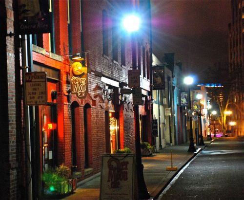 A dimly lit street at night, featuring brick buildings and illuminated signs for bars and cafes.