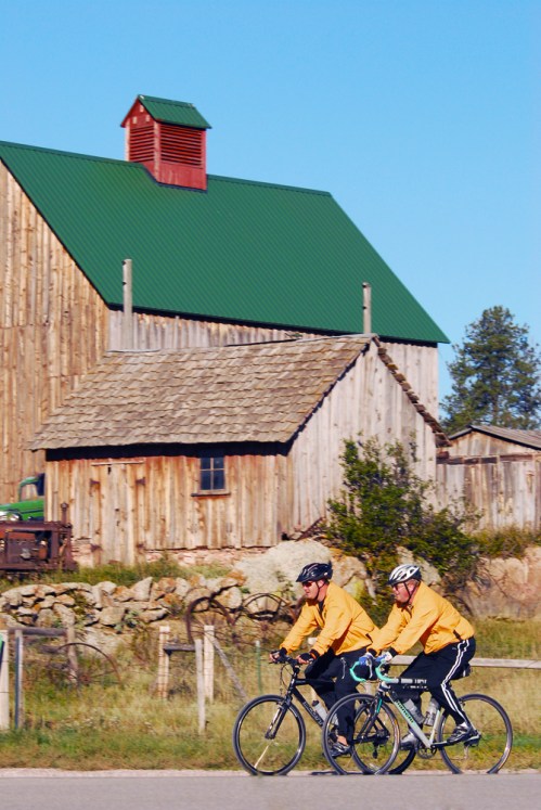 Two cyclists in yellow jackets ride past a rustic wooden barn with a green roof under a clear blue sky.