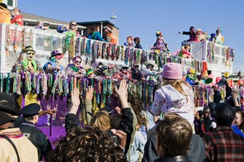 A lively parade scene with colorful floats, performers in costumes, and a crowd celebrating with raised hands and beads.