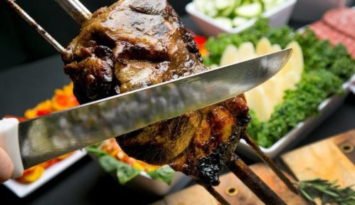 A close-up of a knife slicing through grilled meat on a skewer, with fresh vegetables in the background.