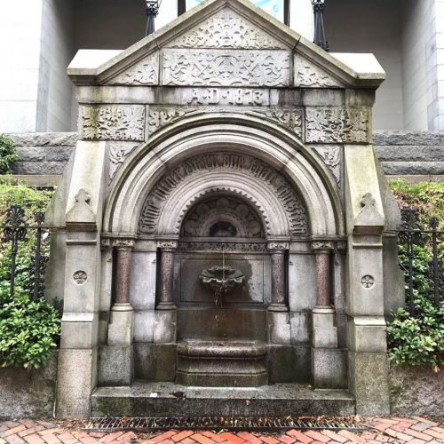Historic stone fountain with intricate carvings, surrounded by greenery and a decorative iron fence.