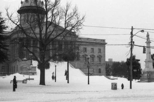 A snowy scene featuring a historic building, a tree, and a statue, with people walking in the winter landscape.