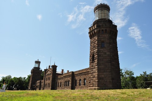 Historic stone lighthouse towers against a blue sky, surrounded by green grass and trees.