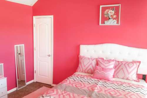 A bright pink bedroom featuring a white bed with patterned pillows, a mirror, and a floral wall art.