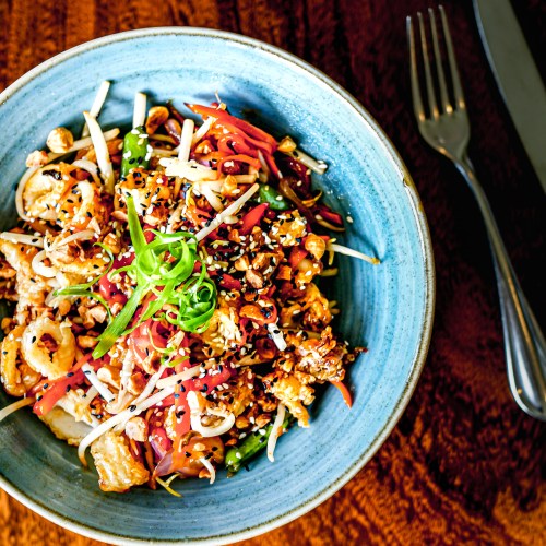 A colorful bowl of mixed salad with vegetables, nuts, and sesame seeds, served on a wooden table with cutlery.