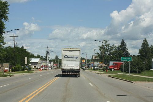 A view of a road with a truck in the foreground and a sign for Antigo in the distance under a partly cloudy sky.