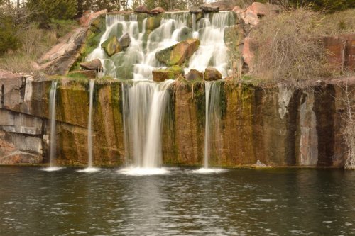 A serene waterfall cascading over rocky ledges into a calm pool, surrounded by greenery and natural stone.