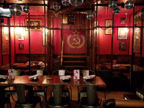 A vibrant restaurant interior with red walls, decorative mirrors, and a Soviet emblem, featuring wooden tables and chairs.