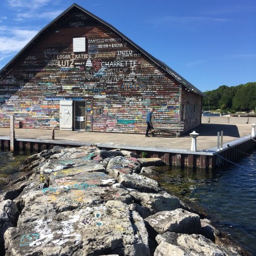 A colorful, graffiti-covered barn by the water, with a person walking along a rocky pier under a clear blue sky.