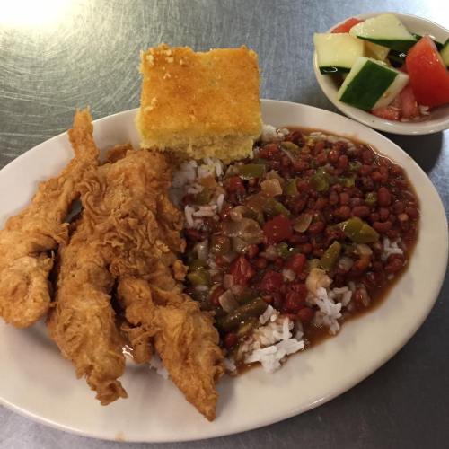 A plate of fried chicken strips, rice with red beans, cornbread, and a side of fresh fruit salad.
