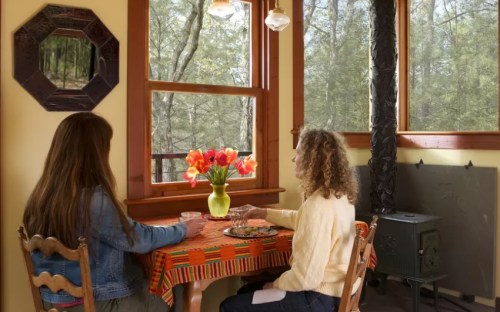 Two people sit at a colorful table by a window, enjoying a view of trees and a vase of flowers.