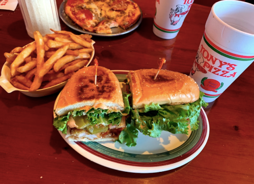 A plate with a sandwich filled with lettuce and pickles, accompanied by a side of fries and a pizza in the background.