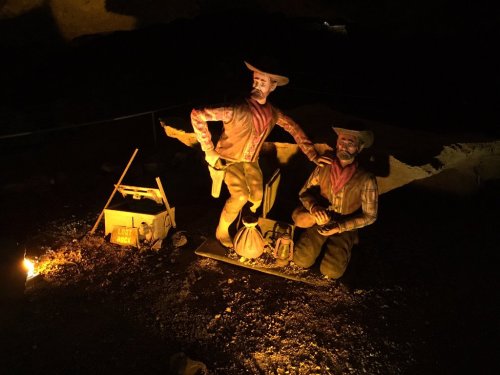 Two figurines of miners in a dark cave, one standing and one kneeling, with mining tools and a candle nearby.