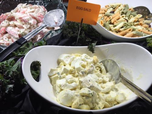 A buffet display featuring egg salad in a white bowl, with pasta salad and shrimp salad in the background.