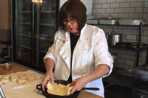 A woman in a chef's coat prepares dough in a kitchen, focused on shaping it in a cast iron skillet.
