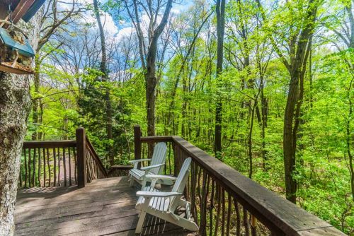 A wooden deck with two white chairs overlooks a lush green forest under a bright blue sky.