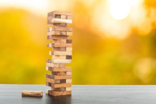 A tall Jenga tower made of wooden blocks, with one block lying on the table, set against a blurred sunset background.