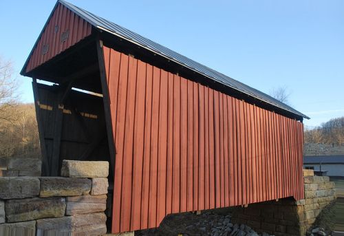 A red covered bridge with a sloped roof, resting on stone supports against a clear blue sky.