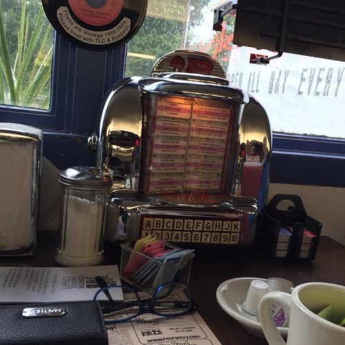 A vintage jukebox sits on a table, surrounded by a sugar container, glasses, and a menu in a cozy diner setting.