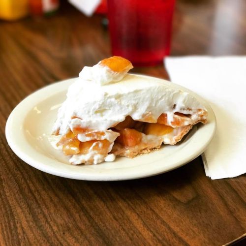 A slice of peach pie topped with whipped cream on a white plate, set on a wooden table.