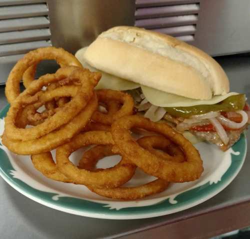 A plate with crispy onion rings and a sandwich filled with peppers, onions, and pickles on a soft bun.