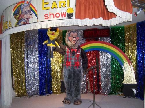 A costumed performer with a wolf mask holds a puppet on stage, decorated with colorful curtains and a rainbow backdrop.