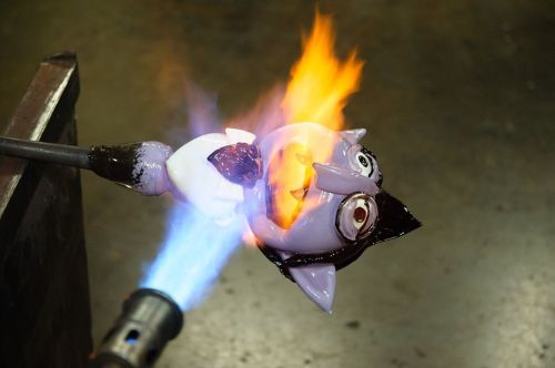 A glass figure being heated with a torch, flames emerging from its body, showcasing vibrant colors and details.