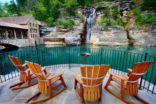 A serene lakeside view with wooden chairs facing a waterfall and rocky cliffs, surrounded by lush greenery.