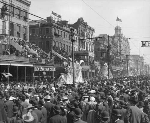 A historic parade scene with large floats and crowds in early 20th-century attire, bustling streets lined with spectators.