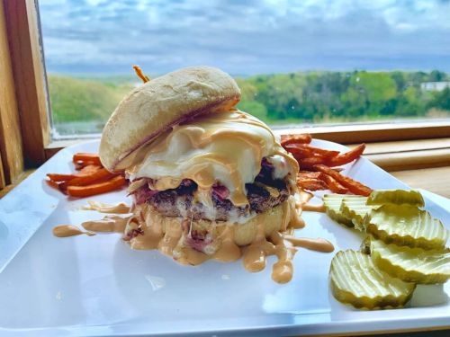 A juicy burger topped with creamy sauce, served with sweet potato fries and pickles, against a scenic window view.