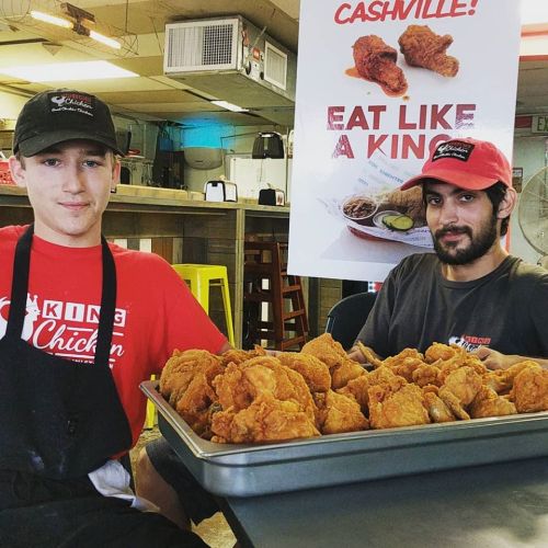 Two young men in restaurant uniforms sit behind a tray of fried chicken, with a promotional sign in the background.