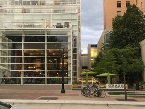 A modern glass building with outdoor seating, bicycles parked nearby, and a sign for a farmer's market stand.