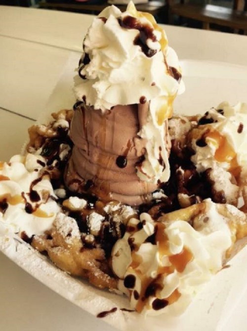 A dessert featuring a chocolate ice cream cone topped with whipped cream, drizzled with chocolate and caramel, on a funnel cake.
