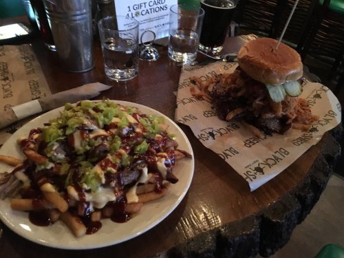 A plate of loaded fries topped with cheese and green onions next to a burger with fried chicken and pickles on a wooden table.