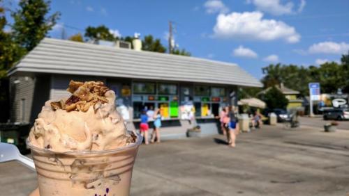 A close-up of a caramel ice cream topped with nuts, with an ice cream shop and people in the background on a sunny day.
