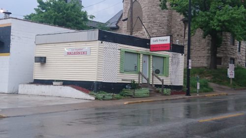 A small, yellow building with green trim, labeled "Café Poland," located on a rainy street.