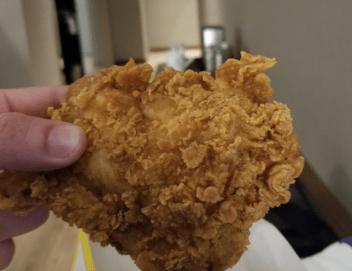 A hand holding a large piece of crispy fried chicken with a golden-brown coating.