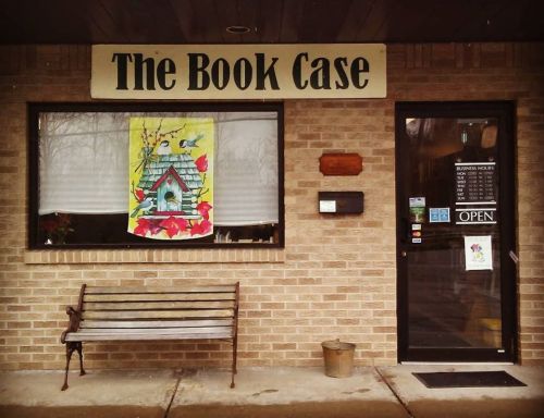 A cozy bookstore entrance with a colorful birdhouse poster, a bench outside, and an "Open" sign on the door.