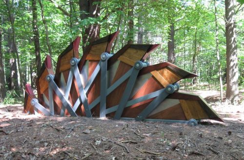 A wooden sculpture resembling a fan, set in a forest with trees and pine needles on the ground.