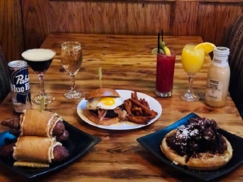 A wooden table with various dishes: burgers, fries, waffles, and drinks including cocktails and beer.