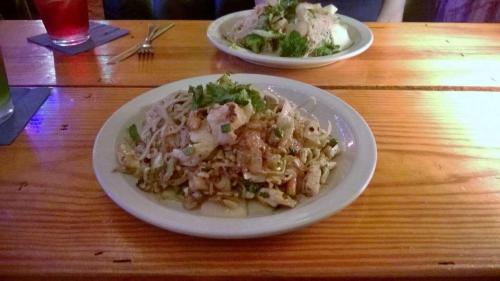 Two plates of Asian-inspired dishes on a wooden table, one with noodles and vegetables, the other with chicken and broccoli.