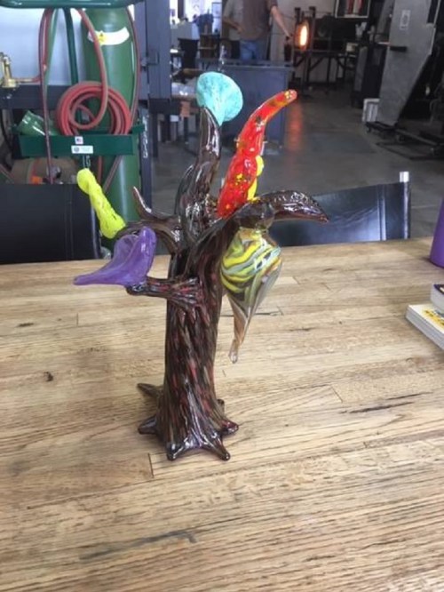A colorful glass sculpture of a tree with various birds perched on its branches, set on a wooden table.