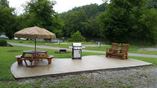 A picnic area with a grill, wooden chairs, and a thatched umbrella by a river, surrounded by trees.