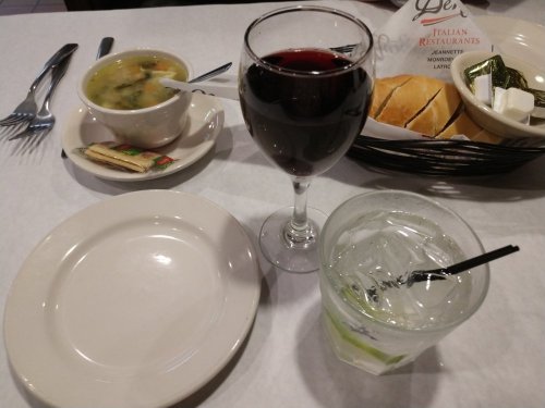 A table setting with a bowl of soup, a glass of red wine, a glass of water with a straw, and a basket of bread rolls.