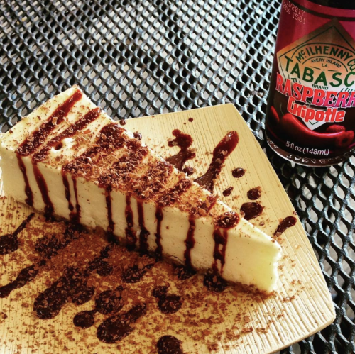 A slice of cheesecake drizzled with chocolate sauce, served on a plate with a can of Tabasco Raspberry Chipotle sauce.