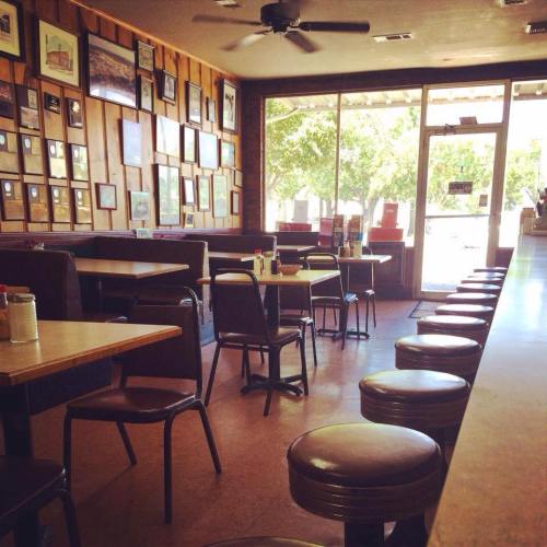 A cozy diner interior with wooden walls, framed photos, tables, and stools, featuring large windows and a welcoming atmosphere.